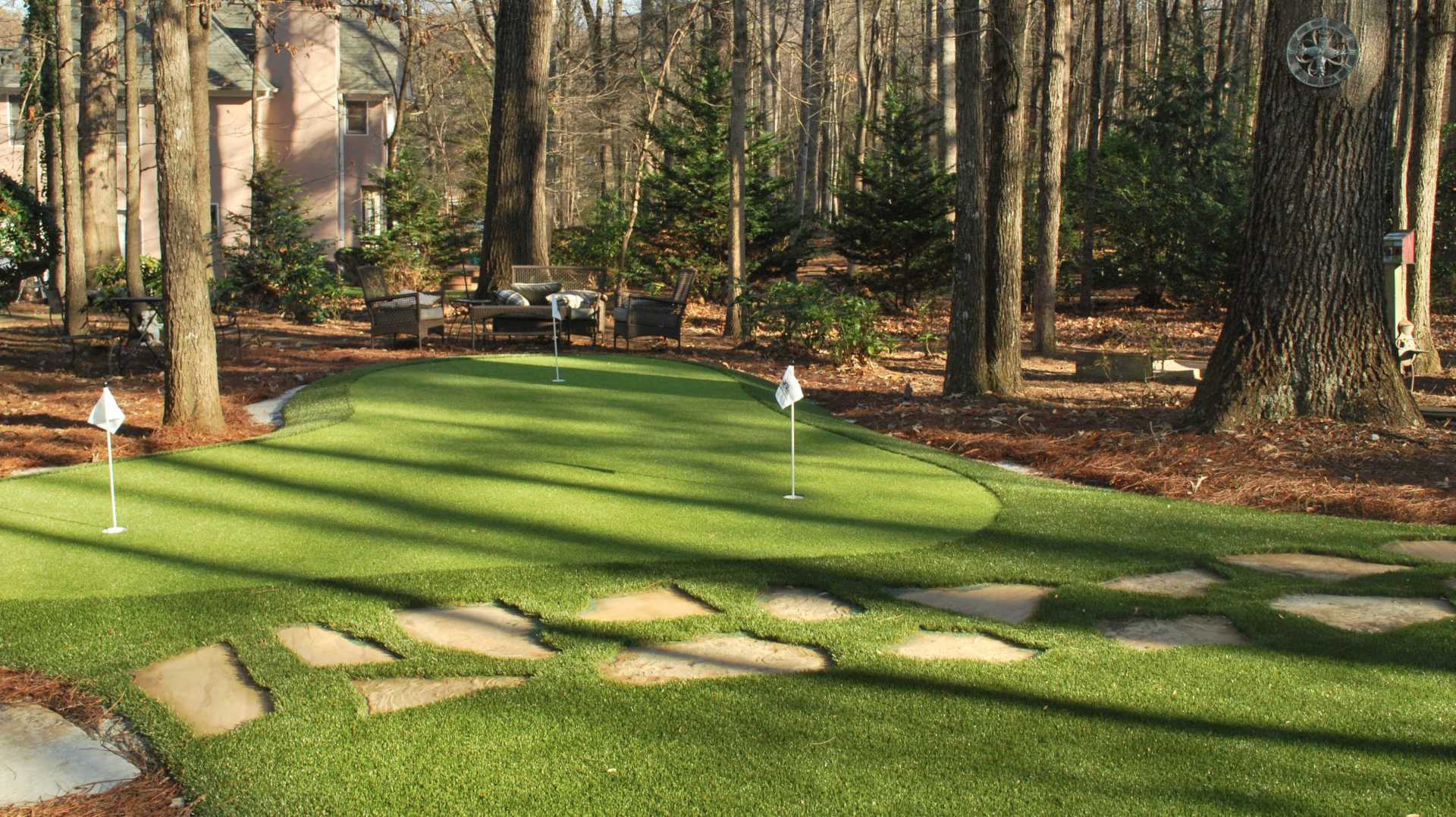Custom backyard putting green with flagsticks, stepping stones, and lounge seating tucked into a wooded residential landscape